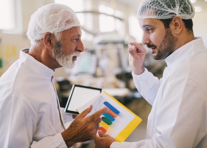 Picture of two male business colleagues in sterile clothes talking about business plans. Standing in bright factory room and discussing about histogram of sales for current month.